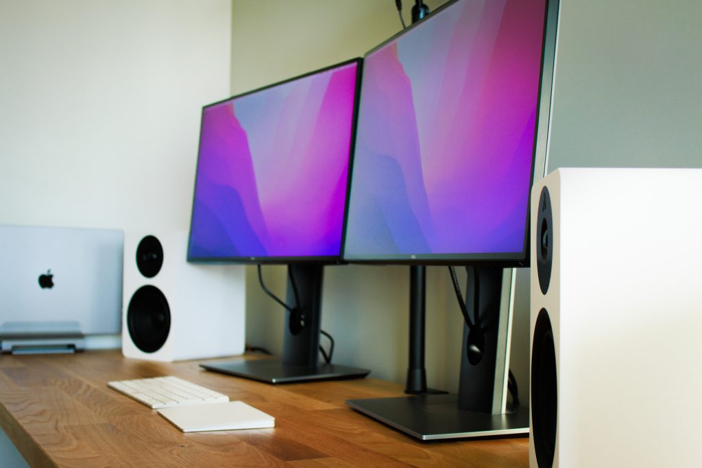 Multi monitor setup with macbook in vertical stand and white speakers on wooden desk